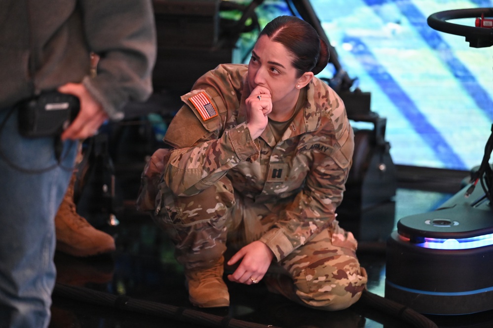 A female U.S. Army officer in OCP (Operational Camouflage Pattern) uniform is crouching and observing what appears to be a military simulator or technology demonstration. She has a captain's rank insignia visible on her uniform and is in a contemplative pose with her hand near her mouth. The setting appears to be an indoor facility with simulation or display equipment visible in the background.
