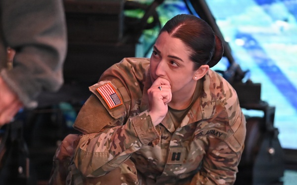 A female U.S. Army officer in OCP (Operational Camouflage Pattern) uniform is crouching and observing what appears to be a military simulator or technology demonstration. She has a captain's rank insignia visible on her uniform and is in a contemplative pose with her hand near her mouth. The setting appears to be an indoor facility with simulation or display equipment visible in the background.