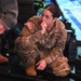 A female U.S. Army officer in OCP (Operational Camouflage Pattern) uniform is crouching and observing what appears to be a military simulator or technology demonstration. She has a captain's rank insignia visible on her uniform and is in a contemplative pose with her hand near her mouth. The setting appears to be an indoor facility with simulation or display equipment visible in the background.