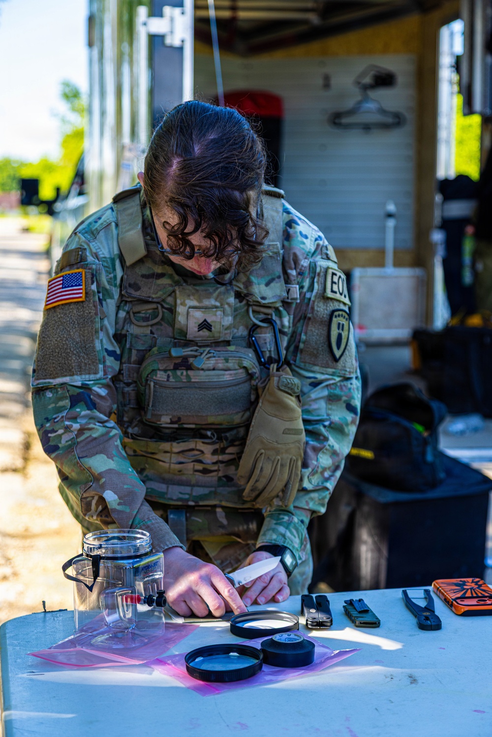 A U.S. Army EOD (Explosive Ordnance Disposal) Sergeant is leaning over a table examining items laid out on a pink surface. The soldier is wearing MultiCam camouflage uniform with a tactical vest, an American flag patch, EOD patch, and a unit shield patch. The setting appears to be an outdoor work area near a building.