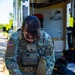 A U.S. Army EOD (Explosive Ordnance Disposal) Sergeant is leaning over a table examining items laid out on a pink surface. The soldier is wearing MultiCam camouflage uniform with a tactical vest, an American flag patch, EOD patch, and a unit shield patch. The setting appears to be an outdoor work area near a building.