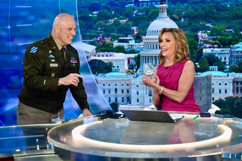 A U.S. Army officer in dress uniform and a woman in a pink dress are seated/standing at a news broadcast desk, smiling and interacting. The woman is holding what appears to be a crystal or glass award or commemorative item. The background displays a large screen showing an aerial view of Washington D.C. with the U.S. Capitol building prominently visible.