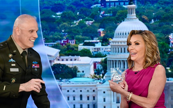 A U.S. Army officer in dress uniform and a woman in a pink dress are seated/standing at a news broadcast desk, smiling and interacting. The woman is holding what appears to be a crystal or glass award or commemorative item. The background displays a large screen showing an aerial view of Washington D.C. with the U.S. Capitol building prominently visible.