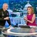 A U.S. Army officer in dress uniform and a woman in a pink dress are seated/standing at a news broadcast desk, smiling and interacting. The woman is holding what appears to be a crystal or glass award or commemorative item. The background displays a large screen showing an aerial view of Washington D.C. with the U.S. Capitol building prominently visible.