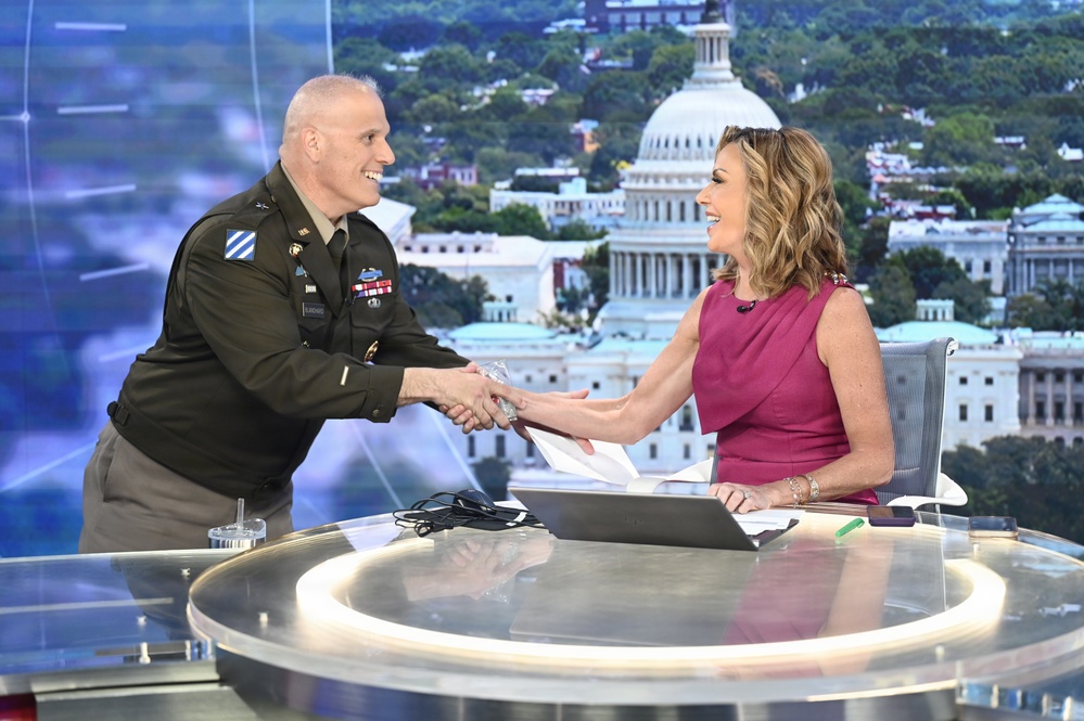 A U.S. Army officer in dress uniform is shaking hands with a female television anchor at a news desk. The setting appears to be a TV news studio with a backdrop displaying the U.S. Capitol building in Washington, D.C. Both individuals are smiling warmly during what appears to be a broadcast segment.