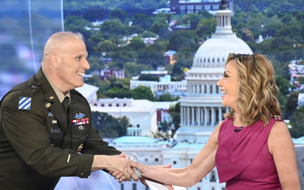 A U.S. Army officer in dress uniform is shaking hands with a female television anchor at a news desk. The setting appears to be a TV news studio with a backdrop displaying the U.S. Capitol building in Washington, D.C. Both individuals are smiling warmly during what appears to be a broadcast segment.