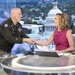 A U.S. Army officer in dress uniform is shaking hands with a female television anchor at a news desk. The setting appears to be a TV news studio with a backdrop displaying the U.S. Capitol building in Washington, D.C. Both individuals are smiling warmly during what appears to be a broadcast segment.