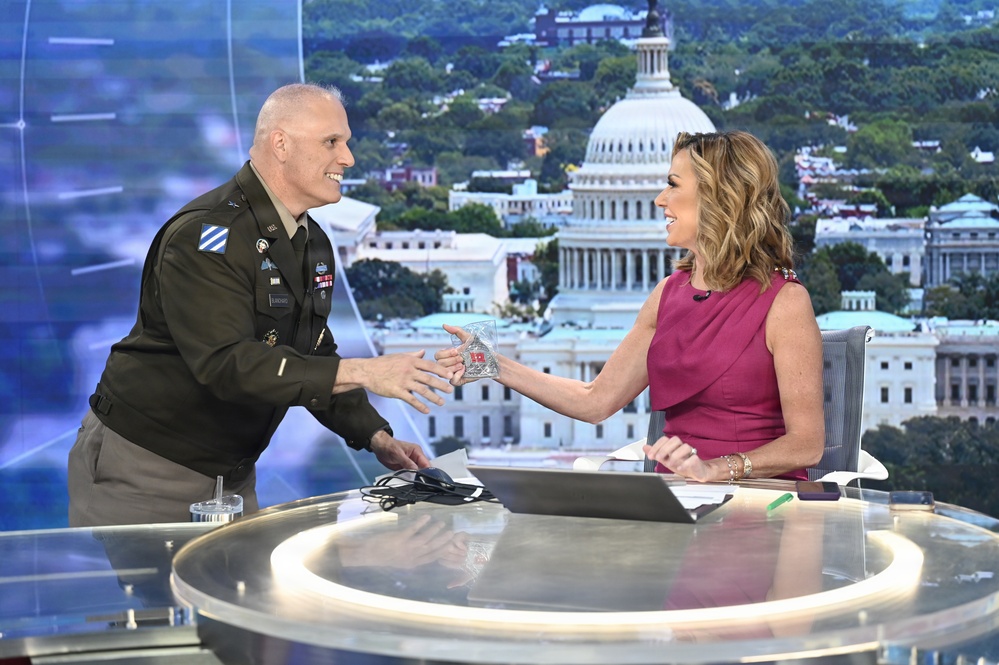 A U.S. Army officer in dress uniform is presenting a glass award or commemorative item to a female television anchor at a news desk. The setting appears to be a television studio with a large backdrop displaying the U.S. Capitol building and the Washington D.C. skyline. Both individuals are smiling and engaged in a positive exchange.