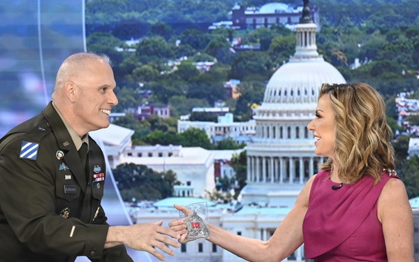A U.S. Army officer in dress uniform is presenting a glass award or commemorative item to a female television anchor at a news desk. The setting appears to be a television studio with a large backdrop displaying the U.S. Capitol building and the Washington D.C. skyline. Both individuals are smiling and engaged in a positive exchange.