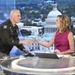 A U.S. Army officer in dress uniform is presenting a glass award or commemorative item to a female television anchor at a news desk. The setting appears to be a television studio with a large backdrop displaying the U.S. Capitol building and the Washington D.C. skyline. Both individuals are smiling and engaged in a positive exchange.