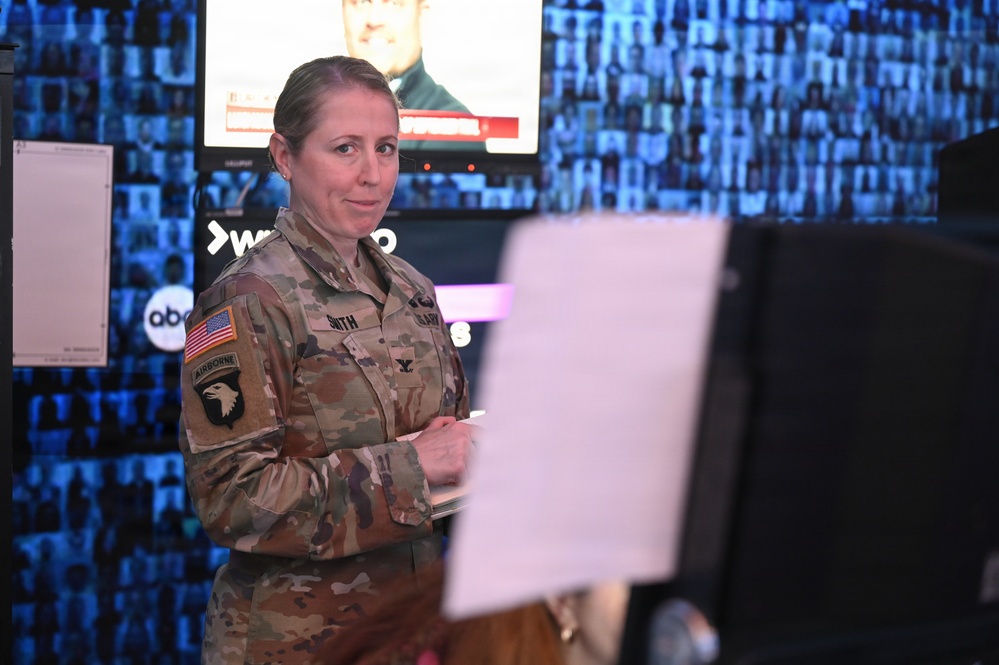 A female U.S. Army soldier in MultiCam (OCP) uniform stands in what appears to be a media or broadcast environment. She is wearing a 101st Airborne Division patch with Airborne tab on her left shoulder and an American flag patch, with a name tape reading 'SMITH'. She appears to be holding papers or a clipboard and looking toward the camera.