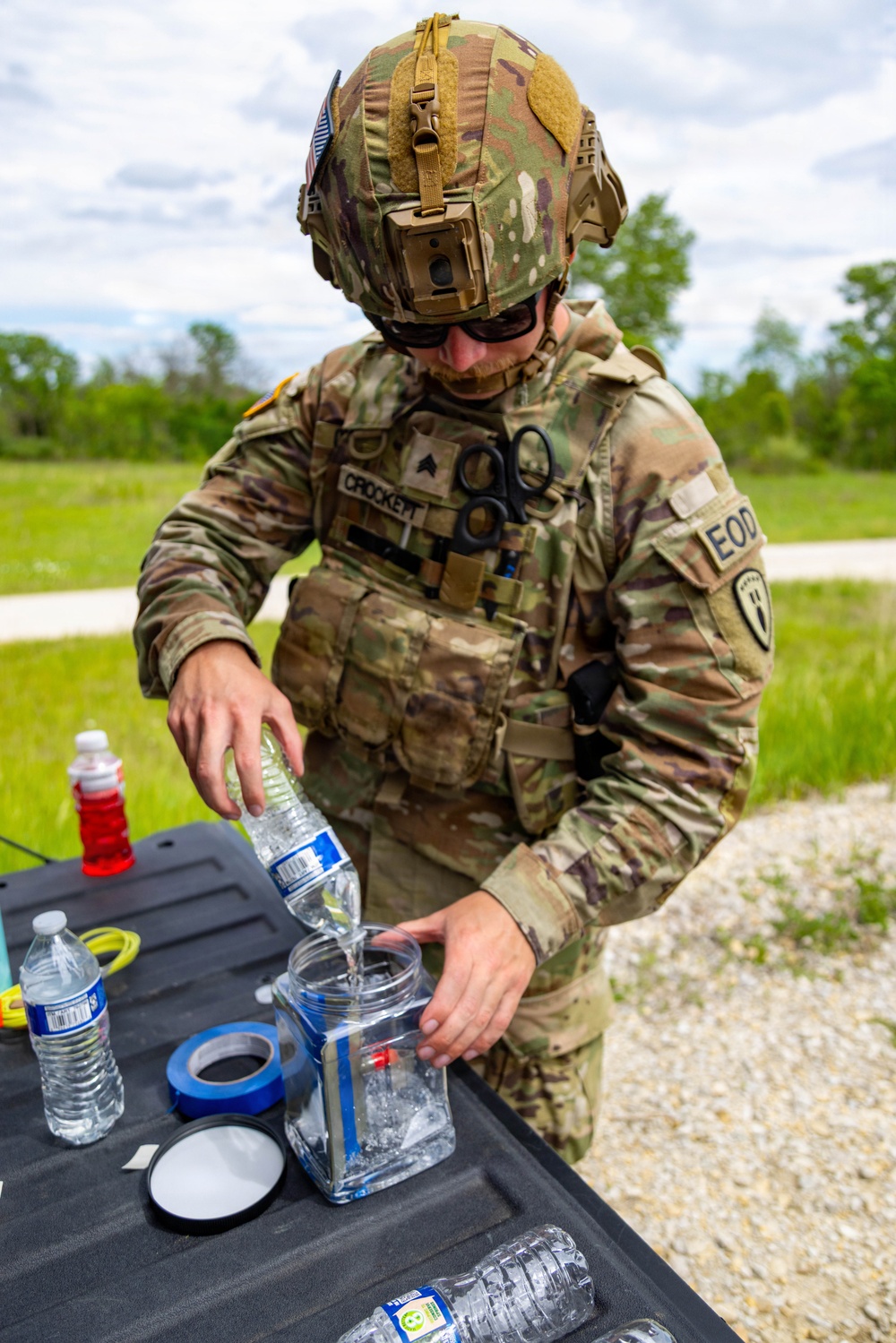 A U.S. Army EOD (Explosive Ordnance Disposal) specialist identified by name tape as 'CROCKETT' is pouring water from a plastic bottle into a clear container at an outdoor field table. The soldier is wearing full OCP (Operational Camouflage Pattern) uniform with a tactical helmet equipped with a camera mount, ballistic eyewear, and a plate carrier vest with scissors attached. The activity appears to be preparation of a water-based tool or device used in EOD operations.