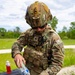 A U.S. Army EOD (Explosive Ordnance Disposal) specialist identified by name tape as 'CROCKETT' is pouring water from a plastic bottle into a clear container at an outdoor field table. The soldier is wearing full OCP (Operational Camouflage Pattern) uniform with a tactical helmet equipped with a camera mount, ballistic eyewear, and a plate carrier vest with scissors attached. The activity appears to be preparation of a water-based tool or device used in EOD operations.