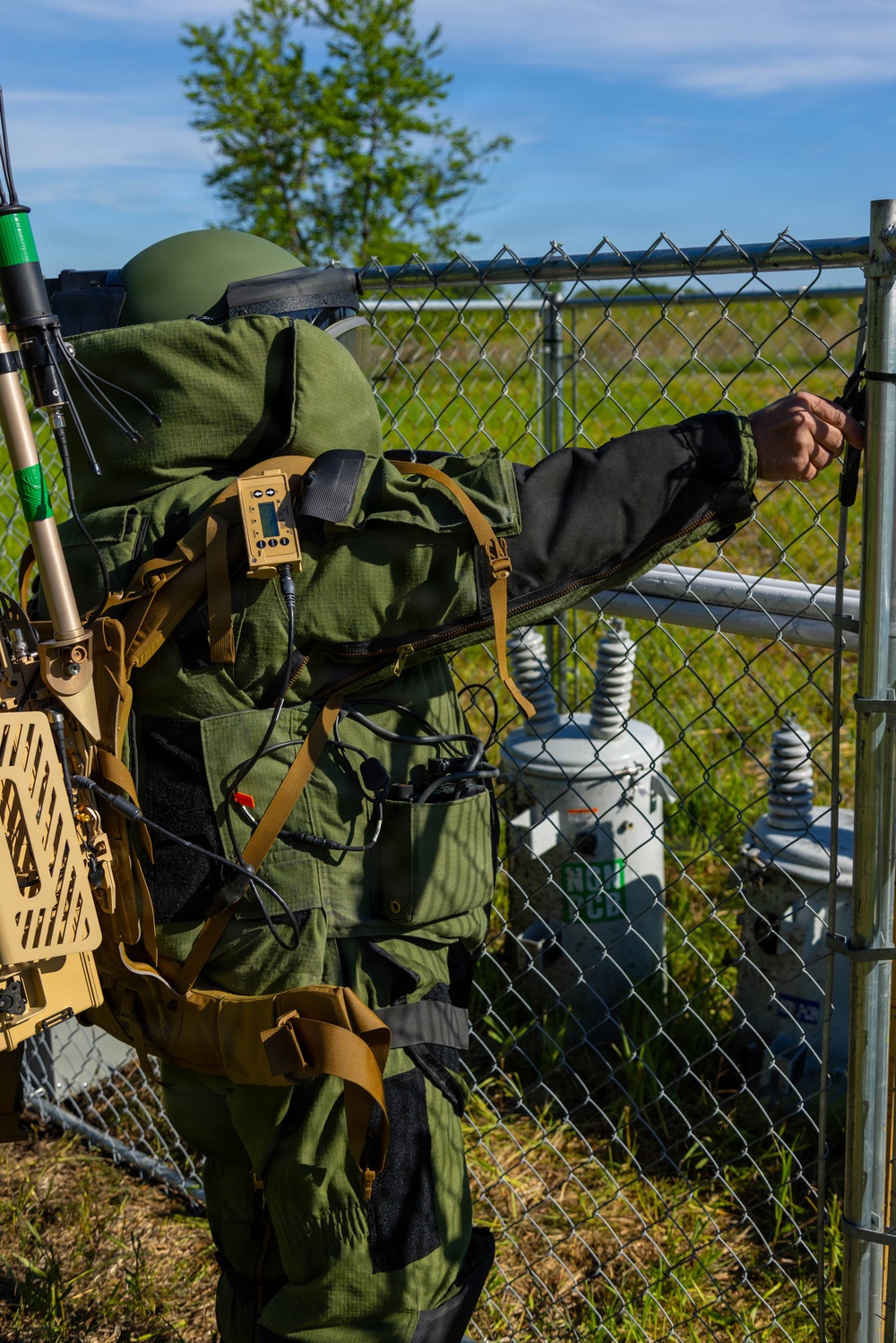 A person wearing an EOD (Explosive Ordnance Disposal) bomb suit in olive green is approaching a chain-link fence gate, reaching toward the latch. The individual is equipped with a helmet, blast-resistant suit, and a tan-colored electronic backpack system with antennas, consistent with EOD or CBRN protective gear. Behind the fence, electrical transformer equipment is visible, suggesting this may be a training exercise at a utility or infrastructure site.
