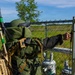 A person wearing an EOD (Explosive Ordnance Disposal) bomb suit in olive green is approaching a chain-link fence gate, reaching toward the latch. The individual is equipped with a helmet, blast-resistant suit, and a tan-colored electronic backpack system with antennas, consistent with EOD or CBRN protective gear. Behind the fence, electrical transformer equipment is visible, suggesting this may be a training exercise at a utility or infrastructure site.