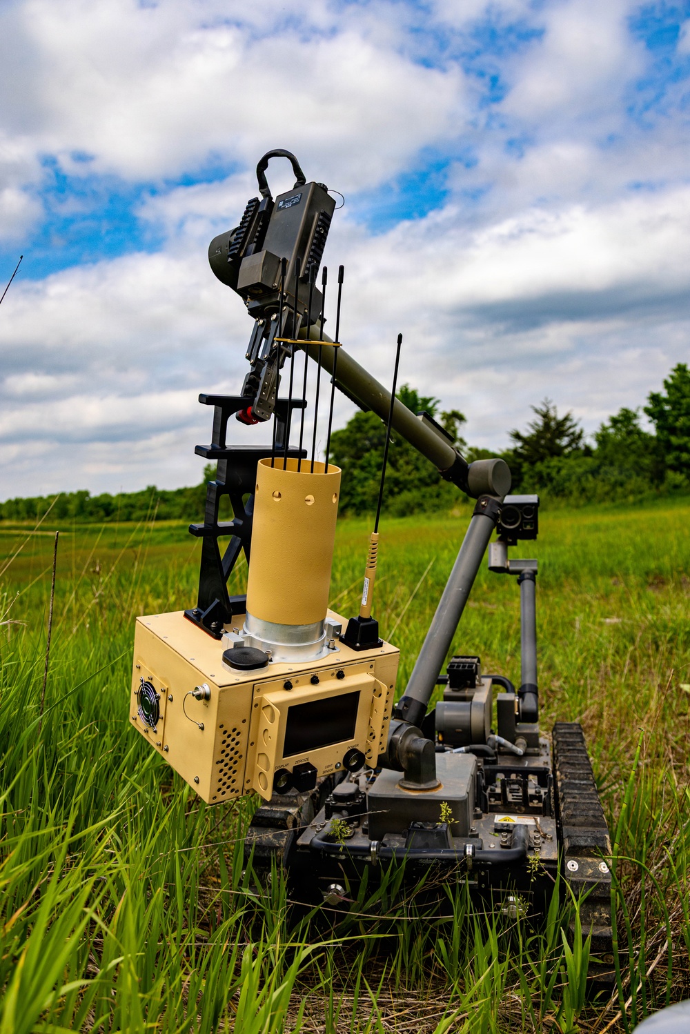 A tracked ground robot is positioned in an open grassy field under a partly cloudy sky. The robot appears to be a military or EOD (Explosive Ordnance Disposal) unmanned ground vehicle (UGV) equipped with a robotic arm, multiple antennas, a cylindrical tube or launcher, and an electronics housing unit. The system appears to be deployed or demonstrated in an outdoor field environment.