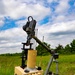A tracked ground robot is positioned in an open grassy field under a partly cloudy sky. The robot appears to be a military or EOD (Explosive Ordnance Disposal) unmanned ground vehicle (UGV) equipped with a robotic arm, multiple antennas, a cylindrical tube or launcher, and an electronics housing unit. The system appears to be deployed or demonstrated in an outdoor field environment.