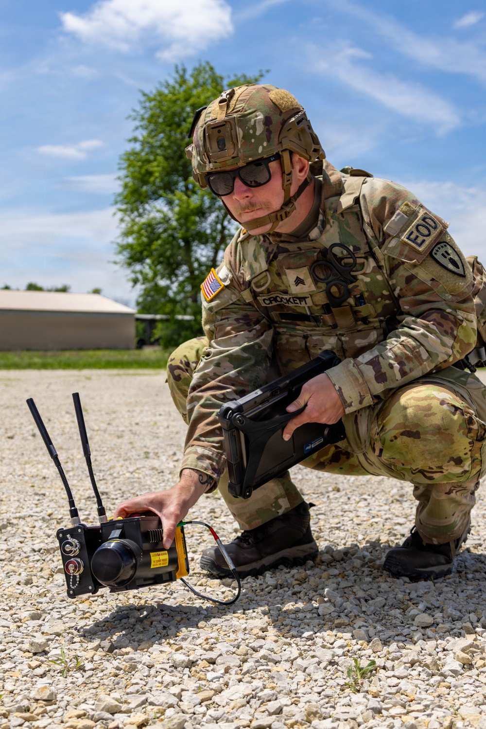 A U.S. Army EOD (Explosive Ordnance Disposal) Sergeant named Crockett is crouching on a gravel surface while operating what appears to be a tactical electronic device with dual antennas. He is holding a ruggedized tablet in one hand while adjusting the ground-placed device with the other. The setting appears to be a military installation or training area during daylight hours.