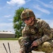 A U.S. Army EOD (Explosive Ordnance Disposal) Sergeant named Crockett is crouching on a gravel surface while operating what appears to be a tactical electronic device with dual antennas. He is holding a ruggedized tablet in one hand while adjusting the ground-placed device with the other. The setting appears to be a military installation or training area during daylight hours.