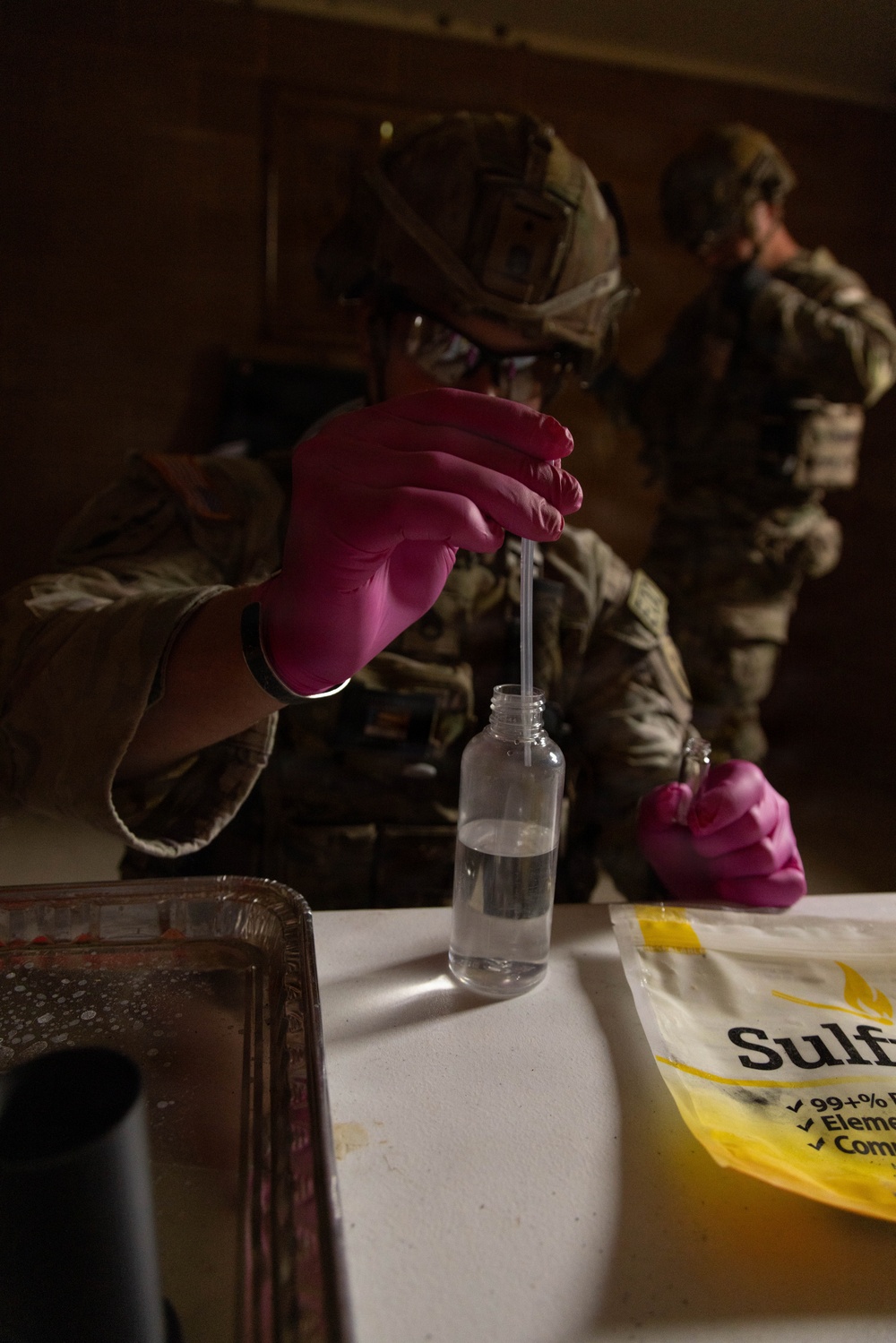 A military personnel wearing combat uniform, helmet, and pink nitrile gloves is conducting what appears to be a chemical or forensic field test, inserting a pipette or test strip into a small clear bottle containing liquid. A yellow bag labeled 'Sulfu...' (likely Sulfur) is visible on the table alongside a metal tray. A second uniformed soldier is visible in the background.