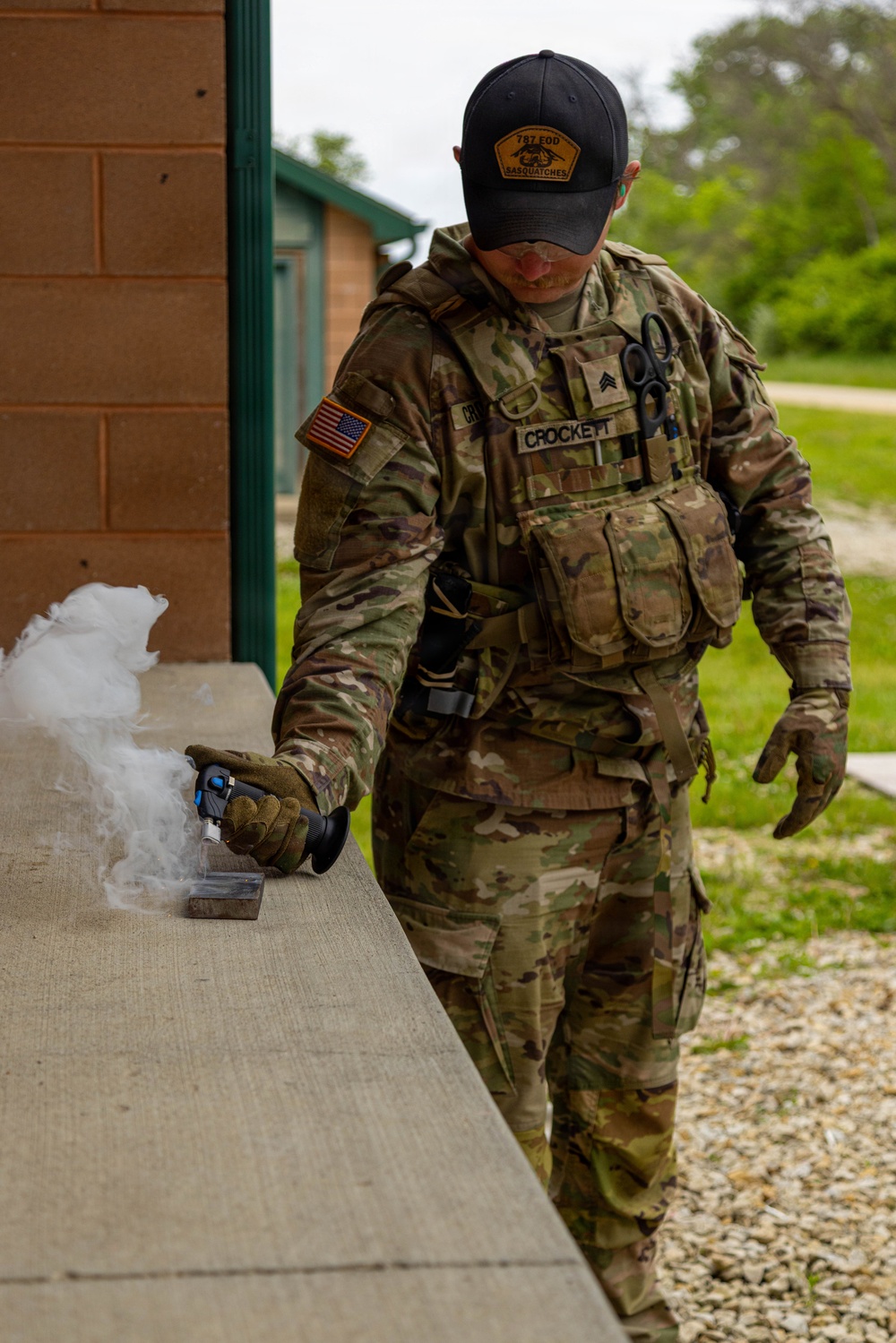 A U.S. Army Sergeant named Crockett, wearing OCP (Operational Camouflage Pattern) uniform with full tactical vest, is applying what appears to be a cryogenic or smoke-producing device to a metal object on a concrete surface. The soldier is assigned to the 787 EOD (Explosive Ordnance Disposal) unit, as indicated by the 'Sasquatches' patch on his cap. The activity appears to be an EOD training or operational procedure conducted outdoors near a building.