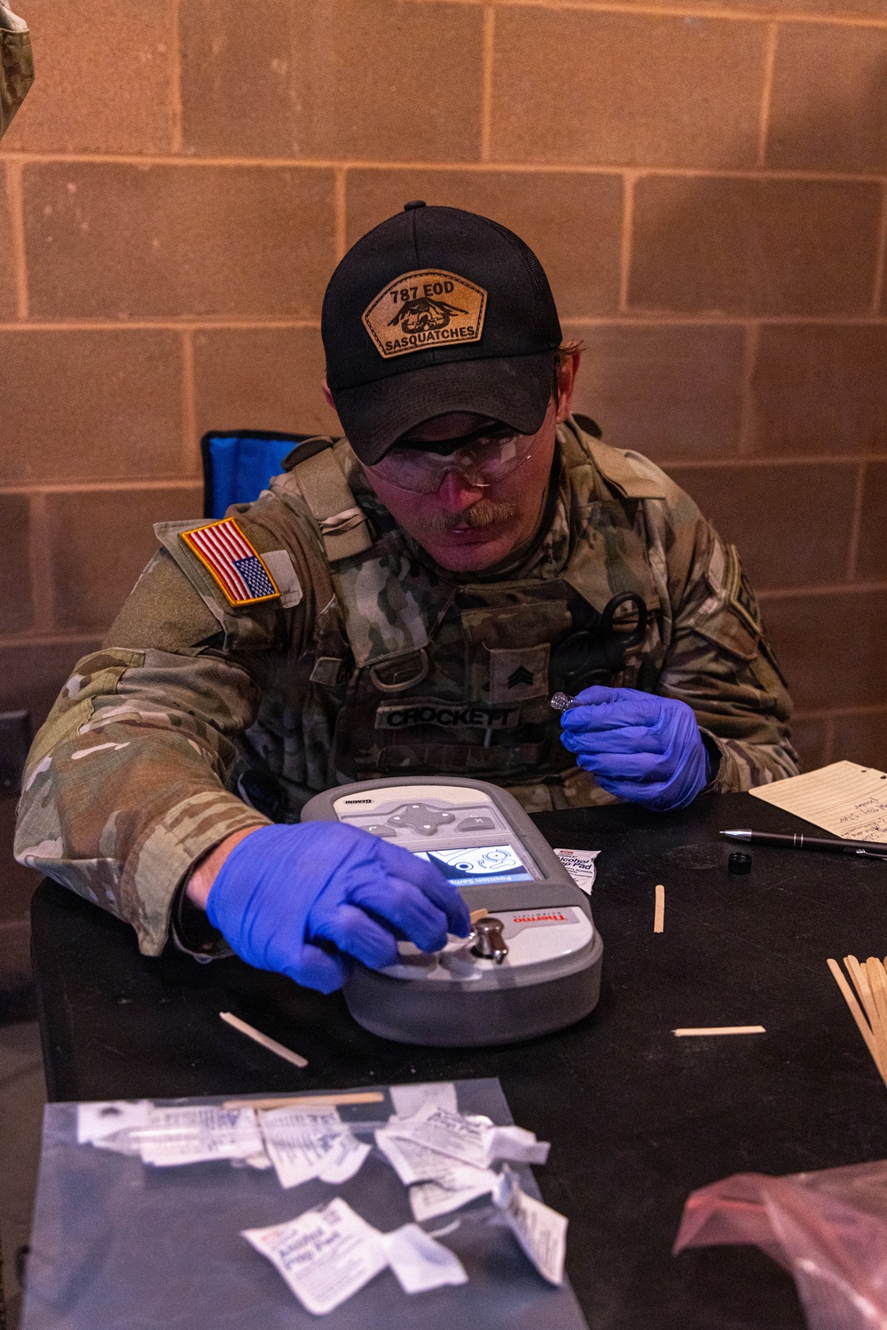 A U.S. Army soldier identified by name tape as 'CROCKETT,' wearing MultiCam uniform with U.S. flag patch and a '787 EOD Sasquatches' unit cap, is operating a Thermo Scientific handheld detection device, likely a TruDefender or similar chemical/explosive analyzer. The soldier is wearing blue nitrile gloves and safety glasses while inserting or manipulating a sample swab into the device. The setting appears to be an indoor training or operational environment with a cinder block wall background.