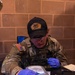 A U.S. Army soldier identified by name tape as 'CROCKETT,' wearing MultiCam uniform with U.S. flag patch and a '787 EOD Sasquatches' unit cap, is operating a Thermo Scientific handheld detection device, likely a TruDefender or similar chemical/explosive analyzer. The soldier is wearing blue nitrile gloves and safety glasses while inserting or manipulating a sample swab into the device. The setting appears to be an indoor training or operational environment with a cinder block wall background.