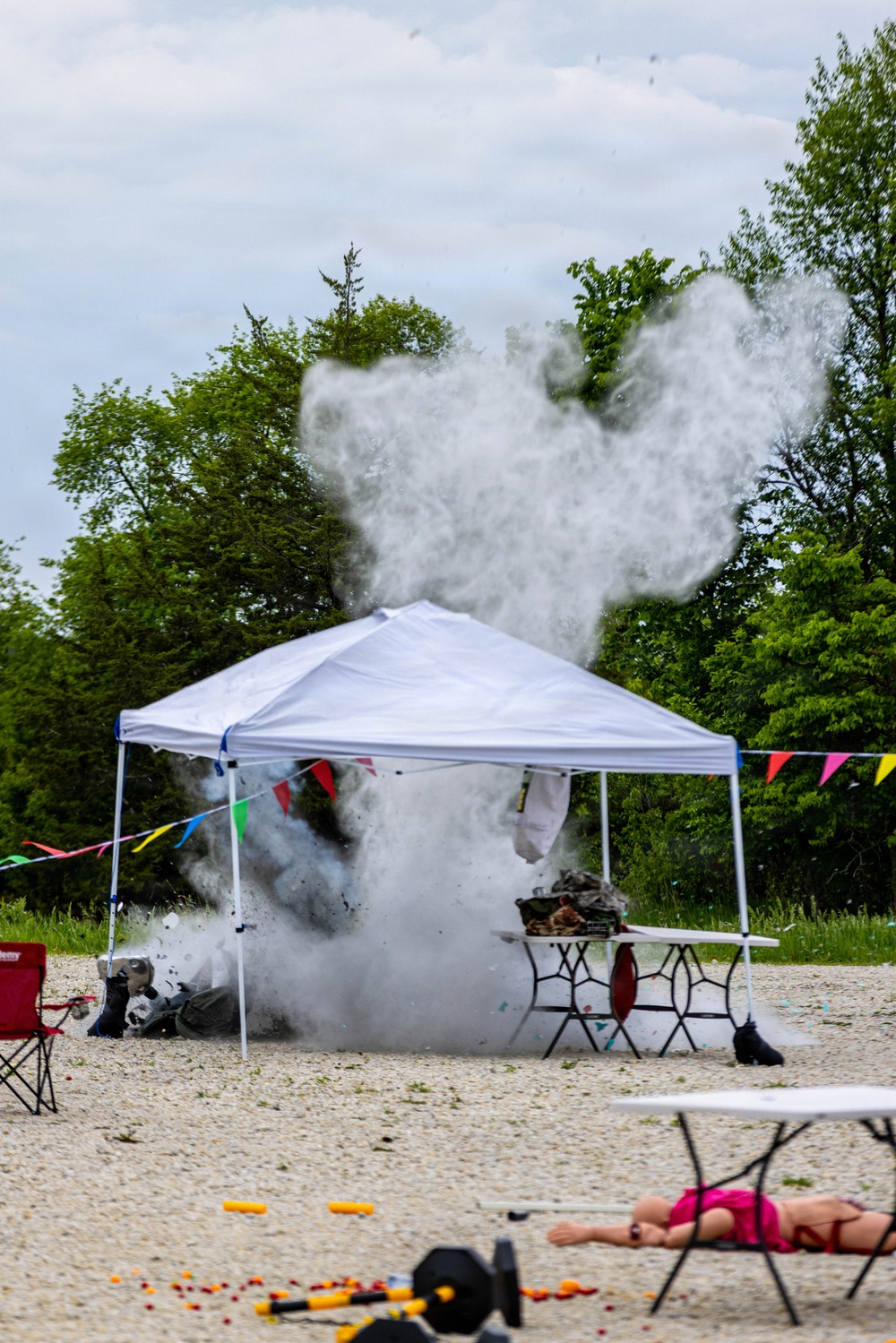 An outdoor explosion or detonation is occurring beneath a white pop-up canopy tent on a gravel/sandy surface, producing a large cloud of white smoke and debris. The scene appears to be a controlled demonstration or event, evidenced by colorful bunting flags, folding chairs, and spectators nearby. A person in pink clothing is visible lying on the ground in the foreground, possibly having taken cover.