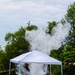 An outdoor explosion or detonation is occurring beneath a white pop-up canopy tent on a gravel/sandy surface, producing a large cloud of white smoke and debris. The scene appears to be a controlled demonstration or event, evidenced by colorful bunting flags, folding chairs, and spectators nearby. A person in pink clothing is visible lying on the ground in the foreground, possibly having taken cover.