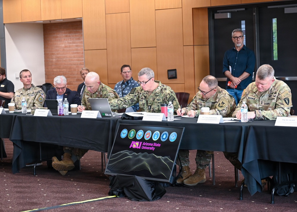 A formal military meeting or briefing is taking place at Arizona State University (ASU), with multiple uniformed military personnel seated at a long black-draped table. The attendees include U.S. Army and U.S. Navy officers, along with civilian participants, engaged in note-taking and reviewing documents. An ASU-branded display sign featuring military branch insignia is prominently placed in the foreground.