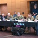 A formal military meeting or briefing is taking place at Arizona State University (ASU), with multiple uniformed military personnel seated at a long black-draped table. The attendees include U.S. Army and U.S. Navy officers, along with civilian participants, engaged in note-taking and reviewing documents. An ASU-branded display sign featuring military branch insignia is prominently placed in the foreground.