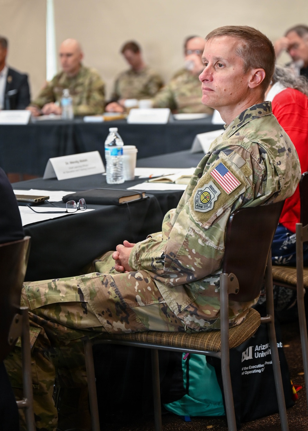 A U.S. military personnel member in OCP (Operational Camouflage Pattern) uniform is seated at a conference or briefing event, appearing to listen attentively. The setting appears to be an academic or institutional meeting, with signage visible indicating Arizona State University. Multiple other military and civilian attendees are visible in the background seated at tables.