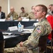 A U.S. military personnel member in OCP (Operational Camouflage Pattern) uniform is seated at a conference or briefing event, appearing to listen attentively. The setting appears to be an academic or institutional meeting, with signage visible indicating Arizona State University. Multiple other military and civilian attendees are visible in the background seated at tables.