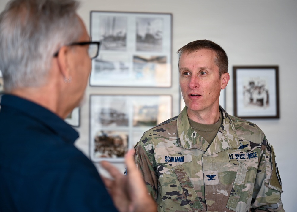 A U.S. Space Force Colonel named Schramm is engaged in conversation with a civilian individual in what appears to be an indoor setting with historical photographs displayed on the wall behind them. The military officer is wearing an OCP (Operational Camouflage Pattern) uniform with a Colonel's eagle insignia and 'U.S. SPACE FORCE' tape visible on his chest. The setting suggests an official visit or briefing environment.
