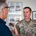 A U.S. Space Force Colonel named Schramm is engaged in conversation with a civilian individual in what appears to be an indoor setting with historical photographs displayed on the wall behind them. The military officer is wearing an OCP (Operational Camouflage Pattern) uniform with a Colonel's eagle insignia and 'U.S. SPACE FORCE' tape visible on his chest. The setting suggests an official visit or briefing environment.