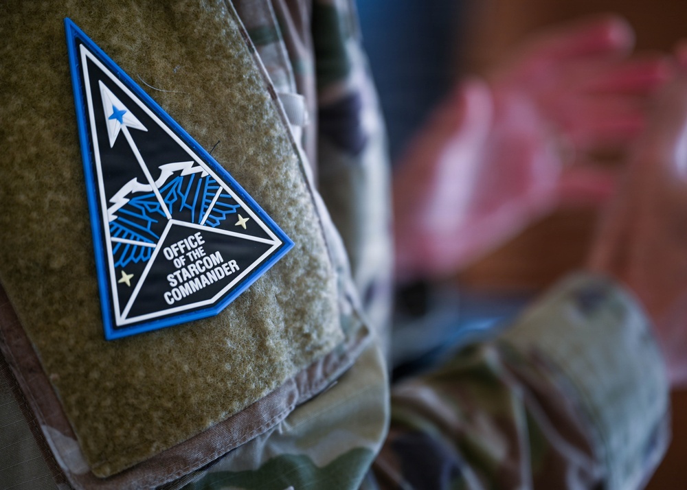 A close-up photograph of a military uniform patch on a camouflage uniform. The patch reads 'Office of the Starcom Commander' and features a triangular design with an eagle emblem, blue border, and stars. A blurred figure in camouflage uniform is visible in the background.