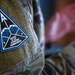 A close-up photograph of a military uniform patch on a camouflage uniform. The patch reads 'Office of the Starcom Commander' and features a triangular design with an eagle emblem, blue border, and stars. A blurred figure in camouflage uniform is visible in the background.
