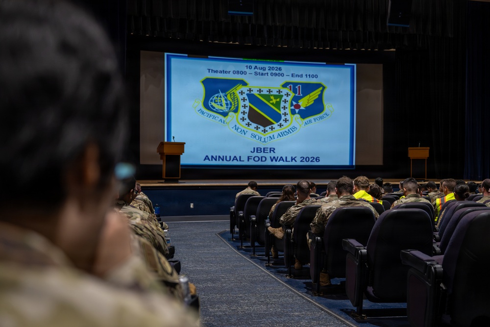 Military personnel are seated in a theater-style auditorium attending a briefing for the JBER Annual FOD Walk 2026, scheduled for 10 August 2026. A large projection screen displays the event details including the Pacific Air Forces and 11th Air Force unit insignia with the motto 'Non Solum Armis.' Attendees are wearing military camouflage uniforms, with some individuals in high-visibility yellow vests visible in the right section.
