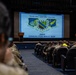 Military personnel are seated in a theater-style auditorium attending a briefing for the JBER Annual FOD Walk 2026, scheduled for 10 August 2026. A large projection screen displays the event details including the Pacific Air Forces and 11th Air Force unit insignia with the motto 'Non Solum Armis.' Attendees are wearing military camouflage uniforms, with some individuals in high-visibility yellow vests visible in the right section.