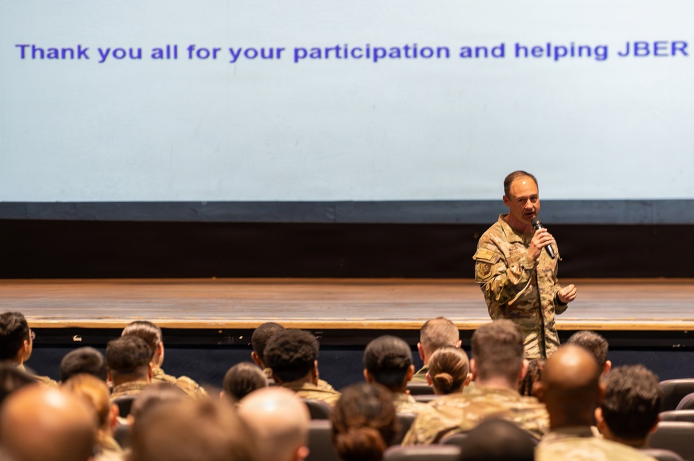 A military officer in camouflage uniform is addressing an audience of uniformed personnel in what appears to be a theater or auditorium setting. A large projection screen behind him displays the message 'Thank you all for your participation and helping JBER,' indicating this is a closing or concluding address at a military event at Joint Base Elmendorf-Richardson (JBER). The speaker is holding a microphone and standing on or near a stage.