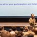 A military officer in camouflage uniform is addressing an audience of uniformed personnel in what appears to be a theater or auditorium setting. A large projection screen behind him displays the message 'Thank you all for your participation and helping JBER,' indicating this is a closing or concluding address at a military event at Joint Base Elmendorf-Richardson (JBER). The speaker is holding a microphone and standing on or near a stage.