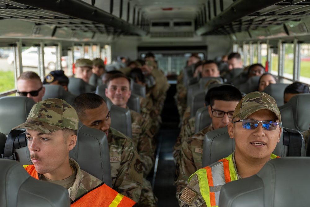 A group of military personnel in OCP (Operational Camouflage Pattern) uniforms are seated aboard a military transport bus. Two individuals in the front seats are wearing high-visibility safety vests, suggesting they may be in supervisory or safety roles. The bus appears fully occupied with service members, likely being transported to or from a duty location.
