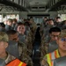 A group of military personnel in OCP (Operational Camouflage Pattern) uniforms are seated aboard a military transport bus. Two individuals in the front seats are wearing high-visibility safety vests, suggesting they may be in supervisory or safety roles. The bus appears fully occupied with service members, likely being transported to or from a duty location.