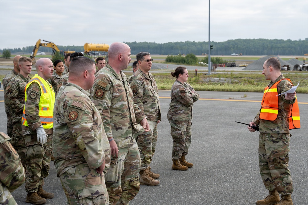 A group of military personnel in OCP (Operational Camouflage Pattern) uniforms are assembled on what appears to be an airfield tarmac. One individual wearing an orange safety vest is addressing the group while holding papers and a radio. The setting appears to be an outdoor briefing or formation at a military airfield with construction equipment visible in the background.