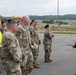 A group of military personnel in OCP (Operational Camouflage Pattern) uniforms are assembled on what appears to be an airfield tarmac. One individual wearing an orange safety vest is addressing the group while holding papers and a radio. The setting appears to be an outdoor briefing or formation at a military airfield with construction equipment visible in the background.