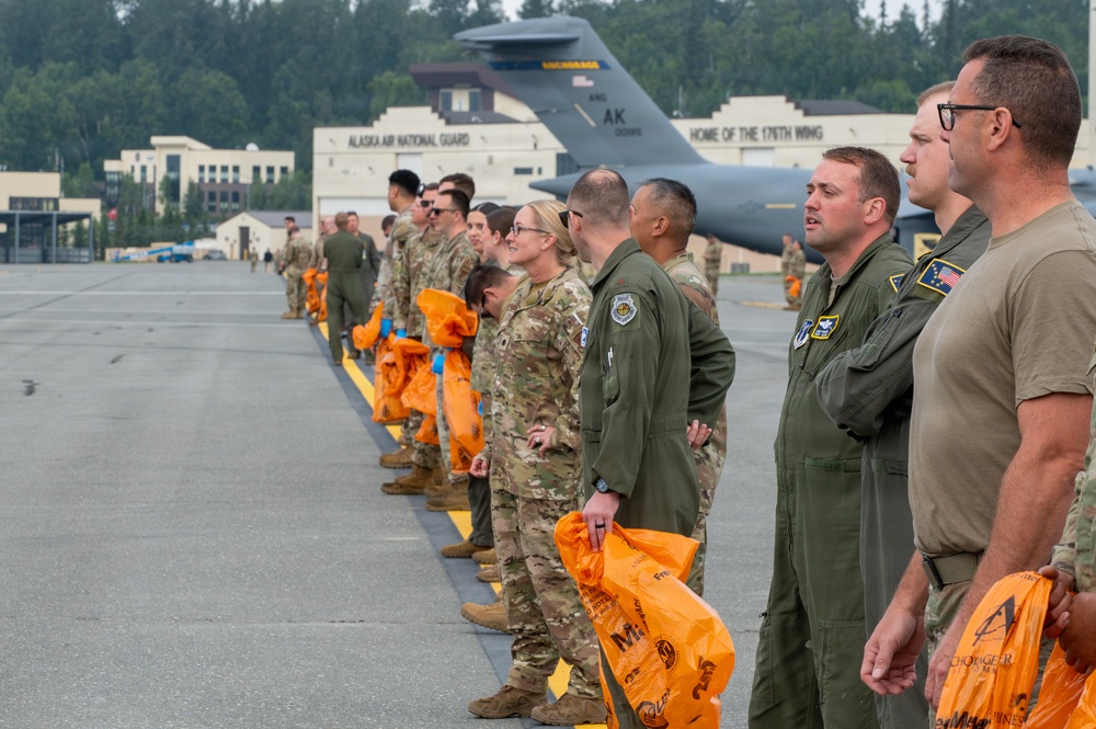 Military personnel are lined up on an airfield tarmac at the Alaska Air National Guard base, home of the 176th Wing, located in Anchorage, Alaska. The group includes both Army soldiers in MultiCam/OCP uniforms and Air Force aircrew in olive green flight suits, many holding orange bags. A large military aircraft, likely a C-17 or KC-135, is visible in the background near the base buildings.