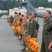 Military personnel are lined up on an airfield tarmac at the Alaska Air National Guard base, home of the 176th Wing, located in Anchorage, Alaska. The group includes both Army soldiers in MultiCam/OCP uniforms and Air Force aircrew in olive green flight suits, many holding orange bags. A large military aircraft, likely a C-17 or KC-135, is visible in the background near the base buildings.