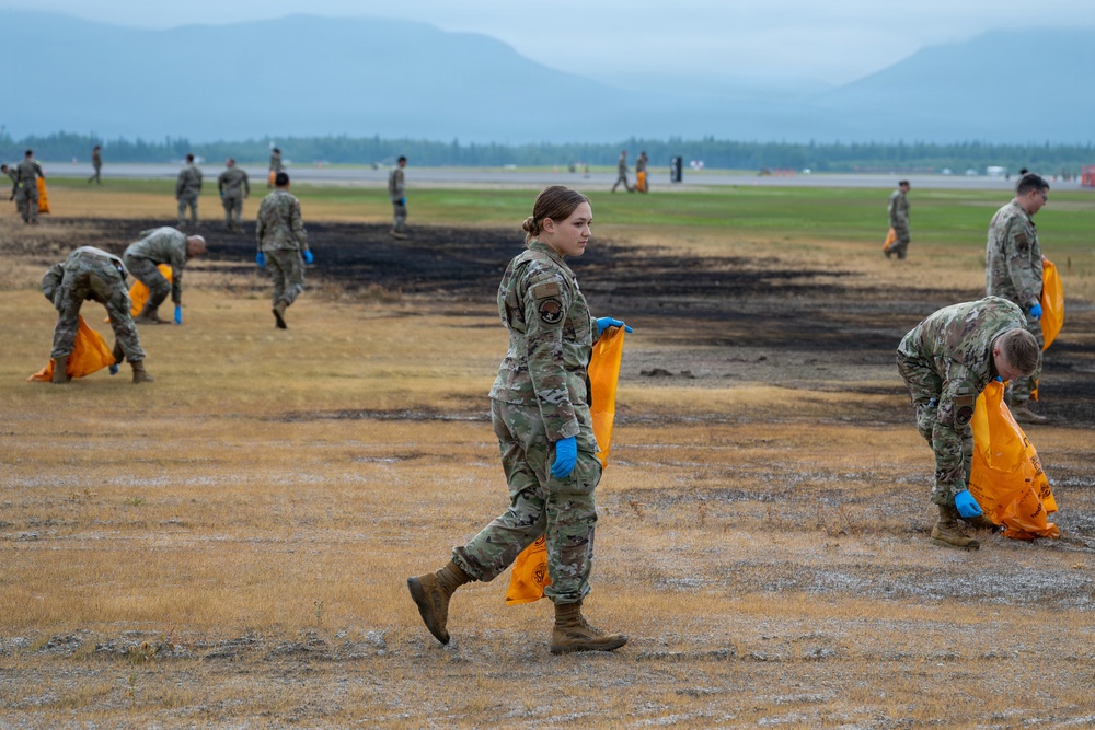 A group of military personnel in camouflage uniforms are conducting a cleanup operation on what appears to be an airfield or military base. They are collecting debris using orange biohazard/waste bags while wearing blue protective gloves. A scorched area of ground is visible, suggesting a recent fire or controlled burn on the field.