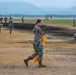 A group of military personnel in camouflage uniforms are conducting a cleanup operation on what appears to be an airfield or military base. They are collecting debris using orange biohazard/waste bags while wearing blue protective gloves. A scorched area of ground is visible, suggesting a recent fire or controlled burn on the field.