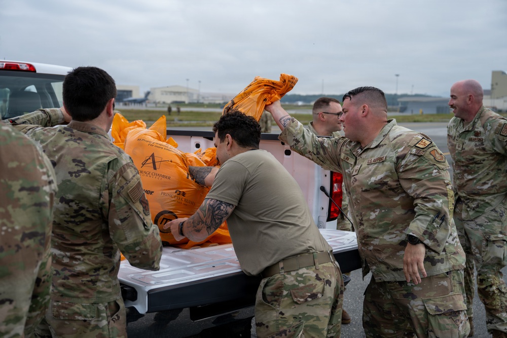 A group of U.S. Air Force personnel in OCP (Operational Camouflage Pattern) uniforms are unloading or loading large orange bags from the bed of a white pickup truck on what appears to be a military airfield. The orange bags are branded with text including 'Storage Chamber' and 'Princess Cruise' logos, suggesting these may be emergency or survival equipment items. The activity appears to be a coordinated logistics or equipment distribution effort on the flight line.